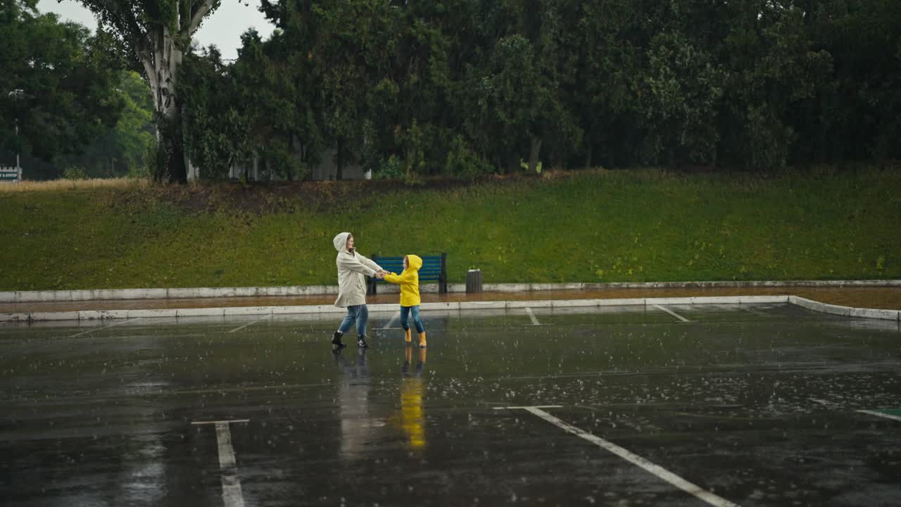 색 재을 입은 행복한 금발 여성과 노란색 재를 입은 십대 소녀가 공원에서 많은 비가 내리는 동안 뛰어내리고 춤을 추고 있습니다.