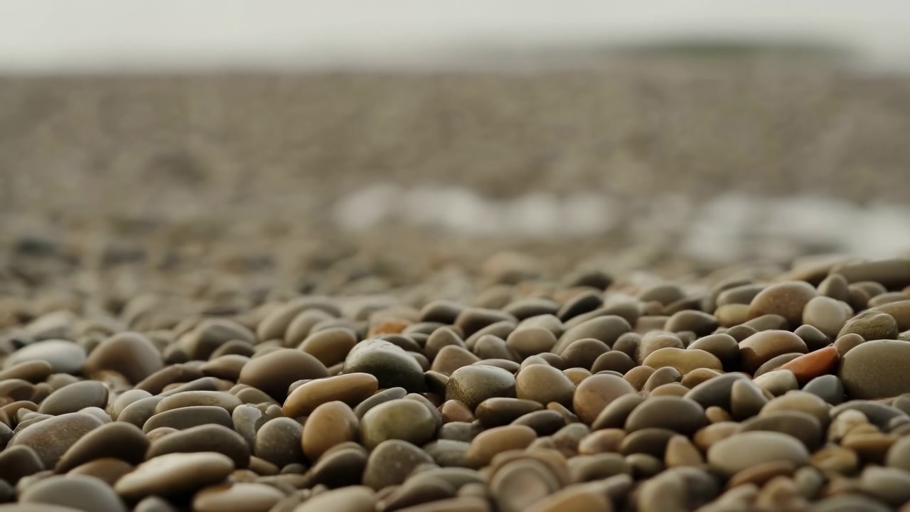 Ciottoli immobili in spiaggia, con ciottoli, sabbia e rocce, texture sabbiosa, da una prospettiva dal basso, con accenti marroni e grigi, e un'atmosfera calma, con luce chiara e composizione equilibrata.