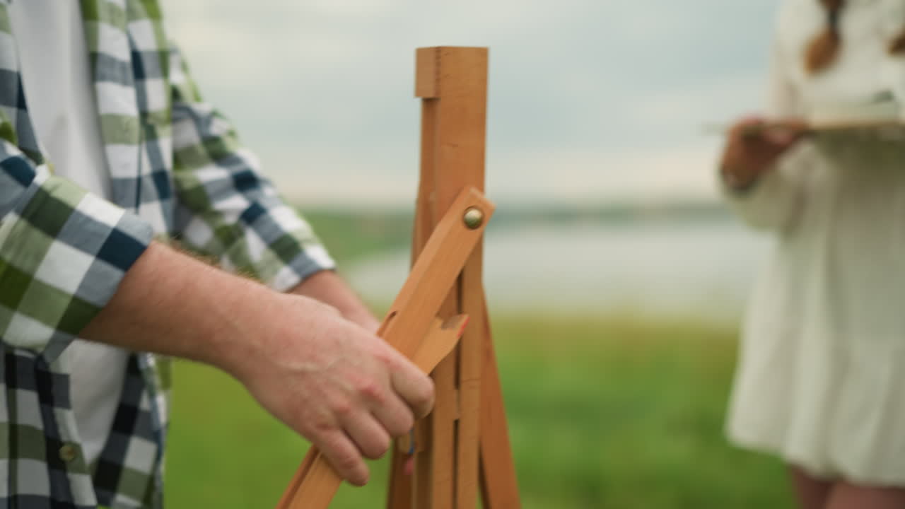 A close-up shot of a man in a plaid shirt holding and setting up a wooden tripod outdoors. In the background, a woman in a white dress is blurred, creating a focus on the man s actions and the tripod
