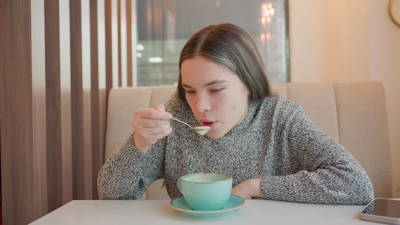 Young lady seated in cafe gently stirs tea with spoon showing satisfaction on face while adjusting herself comfortably on seat in cozy mall environment with warm lights and relaxed ambiance indoors