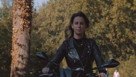 Frontperspektive-Einer-Hübschen-Lächelnden-Europäischen-Jungen-Dame,-Die-An-Einem-Sonnigen-Tag-Ein-Motorrad-Mit-Lederjacke-Im-Wald-Mit-Lebhaften,-Farbenfrohen-Goldenen-Herbstblättern-Fährt