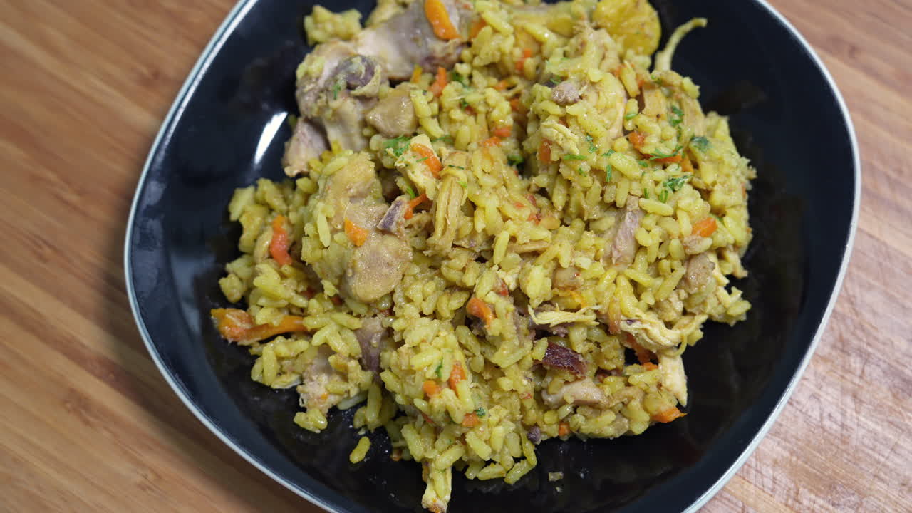 Close-up shot of vibrant Venezuelan Arroz con Pollo on a black plate. The colorful dish is set against a wooden background, highlighting rich textures and aromatic spices under soft lighting