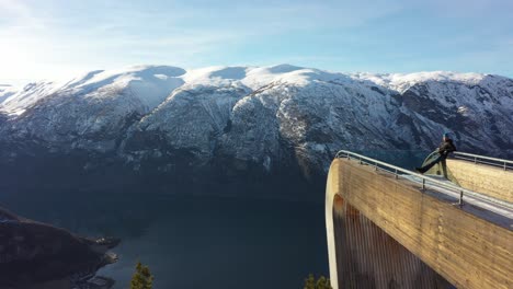 tourist relaxing on tip of stegastein viewpoint platform while drone slowly moving along side and passing edge during golden sunrise