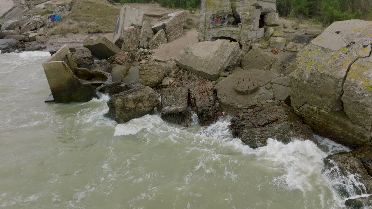 vista aérea de los edificios de fortificación costeros abandonados en los fuertes del norte de karosta, costa del mar báltico, grandes olas, día nublado, cámara lenta, tiro de drones avanzando, inclinado hacia abajo