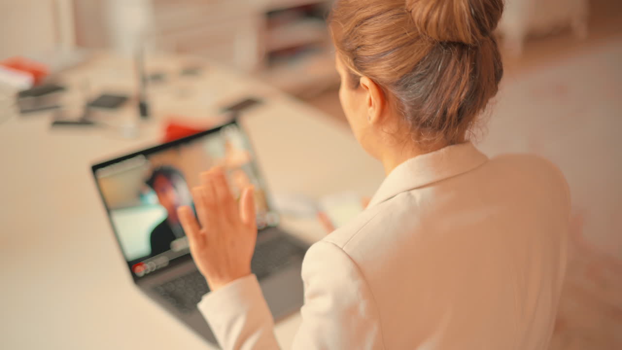 Mujer con blazer blanco hablando en una videoconferencia en un portátil en una mesa mientras sostiene su teléfono