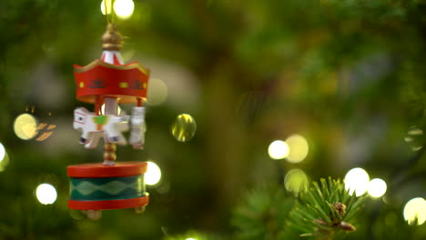 woman hand spinning wooden carousel with white horses as hristmas tree decor hanging on the fir tree with blurred lights in the background in december