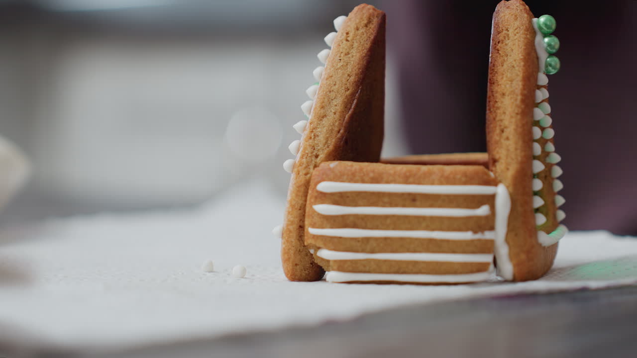 крупный план пекаря, регулирующего имбирные печенье с белой глазурой и декоративными зелеными бусыми, чтобы собрать структуру дома, с слегка размытым фоном