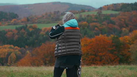 medium shot of a female photographer taking pictures of beautiful autumn natures with bright orange colors with her camera and walking through the field during a windy day in slow motion