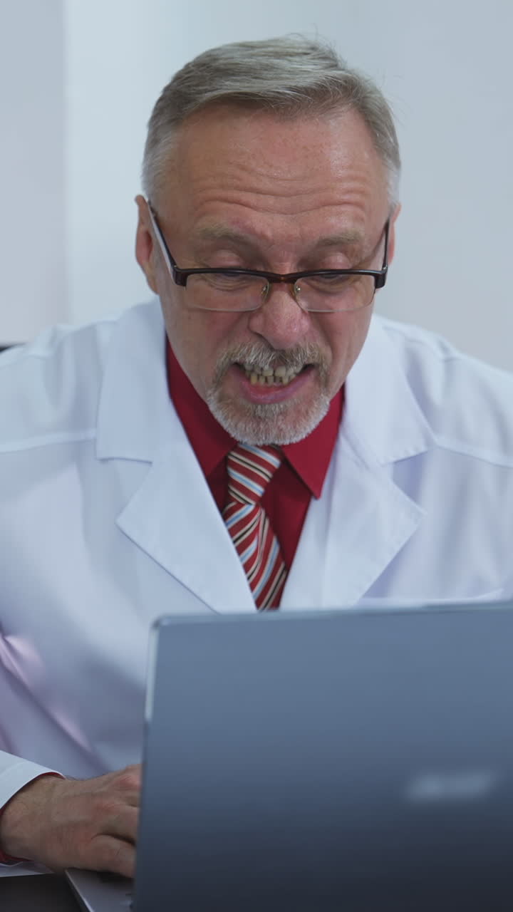 Medical worker having distance video call. Senior doctor in white gown sitting in front of computer and talking to a client in medical center. Online consultation. Vertical video