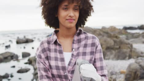 Afroamerikanische-Frau-Lächelt,-Während-Sie-Müll-Auf-Den-Felsen-Am-Meer-Sammelt