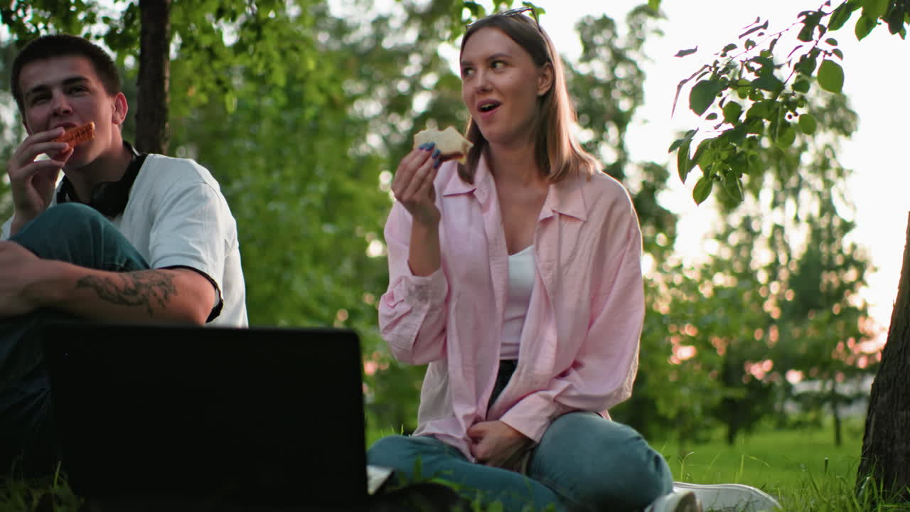 amigos disfrutando de sándwiches a la parrilla al aire libre, una mujer en rosa sentada en la hierba con sándwich en la mano, portátil en el frente, y vegetación en el fondo, disfrutando la comida juntos en un entorno natural