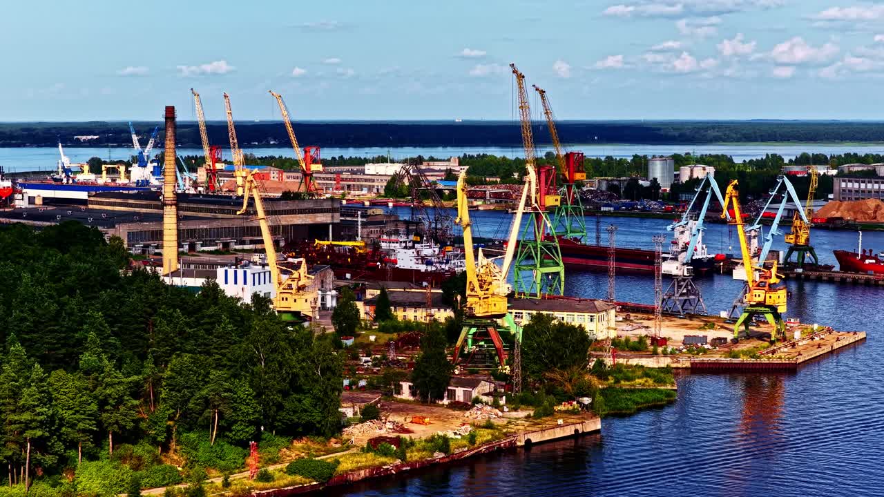 Flygvy över en industrihamn med lastkranar, dockade fartyg, lagerlokaler och skogsklädd strandlinje under en klar sommarhimmel