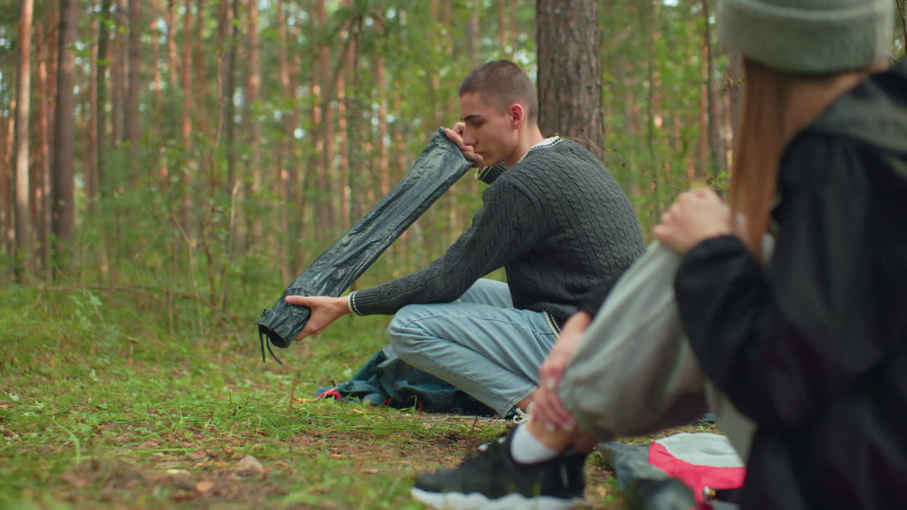 Rear view of lady seated watching boyfriend squat in forest as he carefully unpacks and holds tent pole while preparing campsite surrounded by trees and green environment