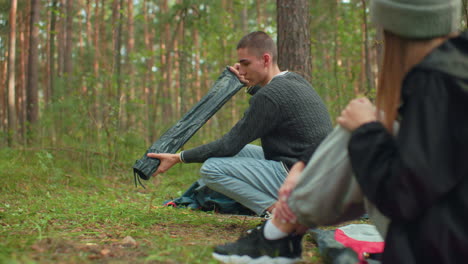rear view of lady seated watching boyfriend squat in forest as he carefully unpacks and holds tent pole while preparing campsite surrounded by trees and green environment