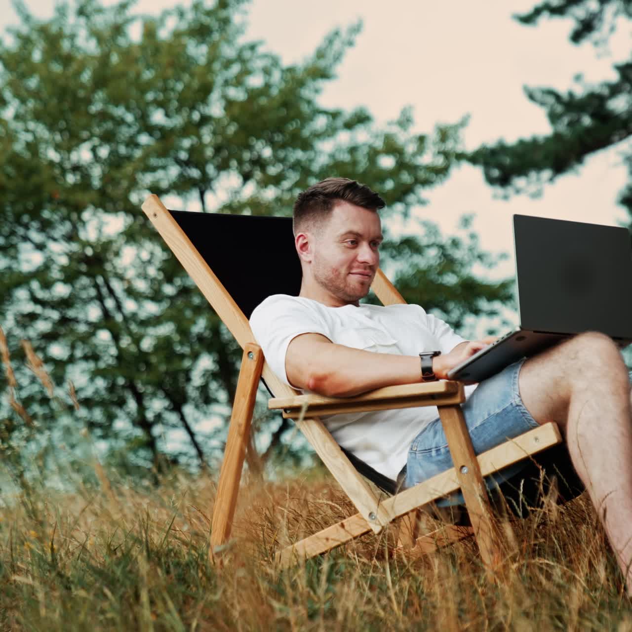 Un uomo freelance sorridente e positivo lavora su una comoda sedia. Lavoro da remoto su Internet immerso nella natura