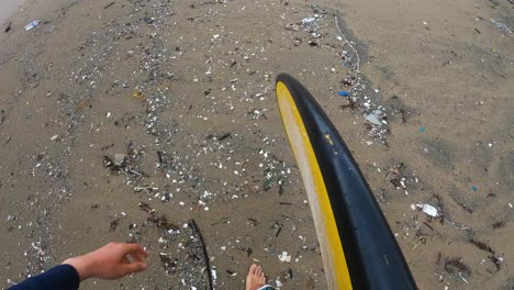 surfer walks along sandy beach covered in micro plastic garbage and other trash