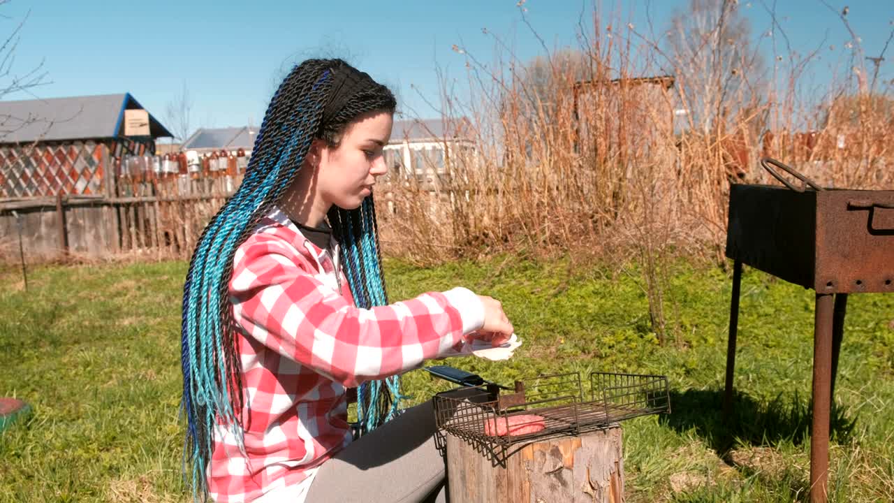 una mujer joven pone chuletas en la parrilla. está con una camisa rosa a cuadros y con el pelo trenzado azul.