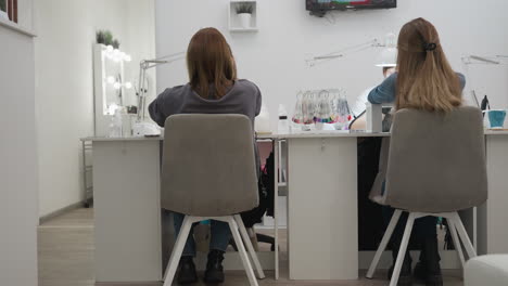 two female customers seated at manicure desks inside well-lit nail salon with technicians out of focus, visible television on wall, nail polish color samples on table, and white interior design
