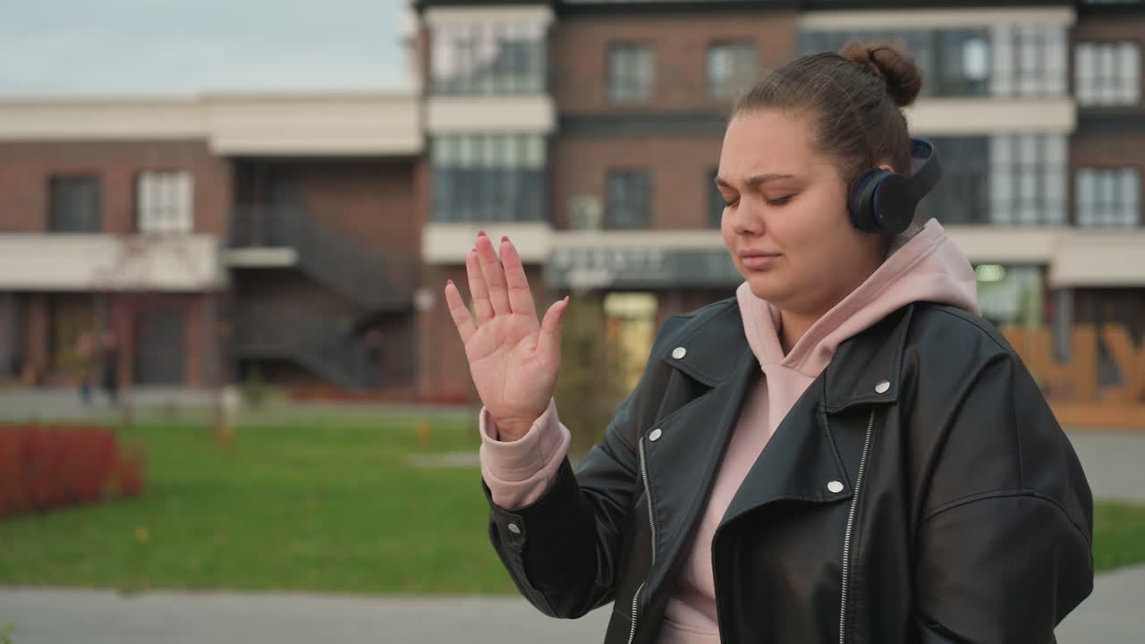 Musician wearing pink hoodie and black leather jacket dances expressively to rhythm of music through headphones moving hand, enjoying beat against backdrop of blurred residential buildings