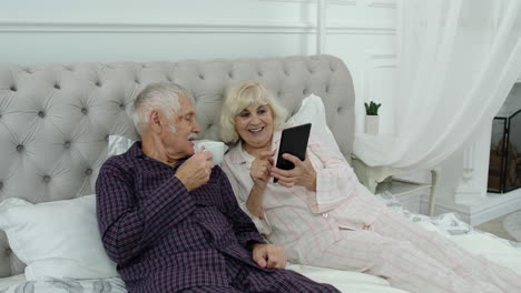 Senior-elderly-couple-wearing-pyjamas-lying-on-bed-looking-on-digital-tablet-making-online-shopping