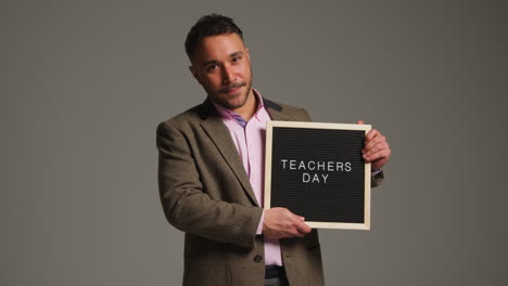 studio portrait of mature male teacher standing against grey background holding notice board reading teachers day