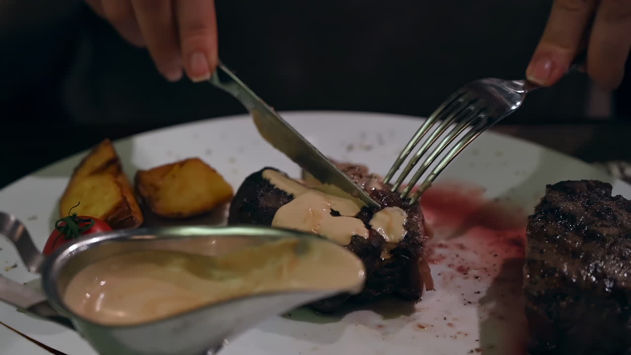 Close up of a woman cutting up a piece of steak with sauce and eating it at a restaurant