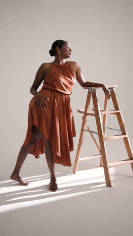 vertical video: elegant woman in stylish dress posing confidently with wooden ladder in studio