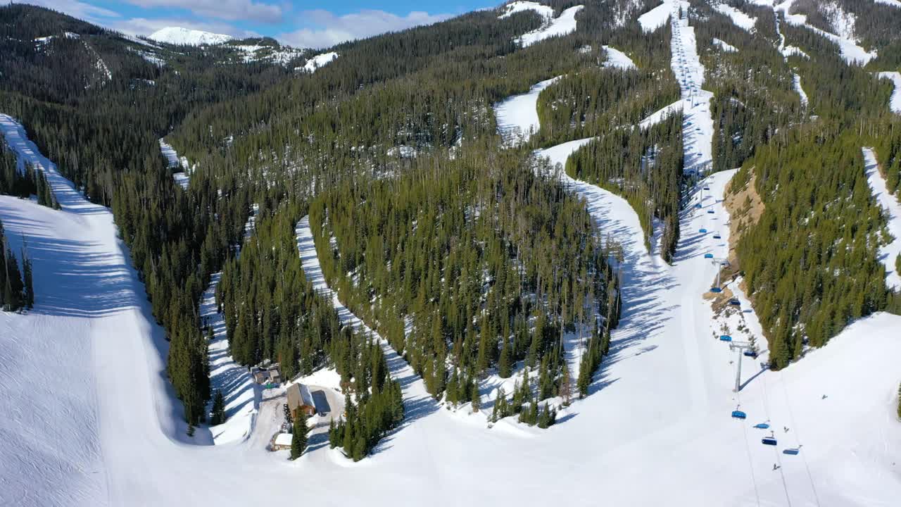Chair lifts full of bundled skiers cross snowy ridges in Big Sky, Montana, captured by drone over icy runs, pine forests, and powder trails beneath a crisp blue winter sky