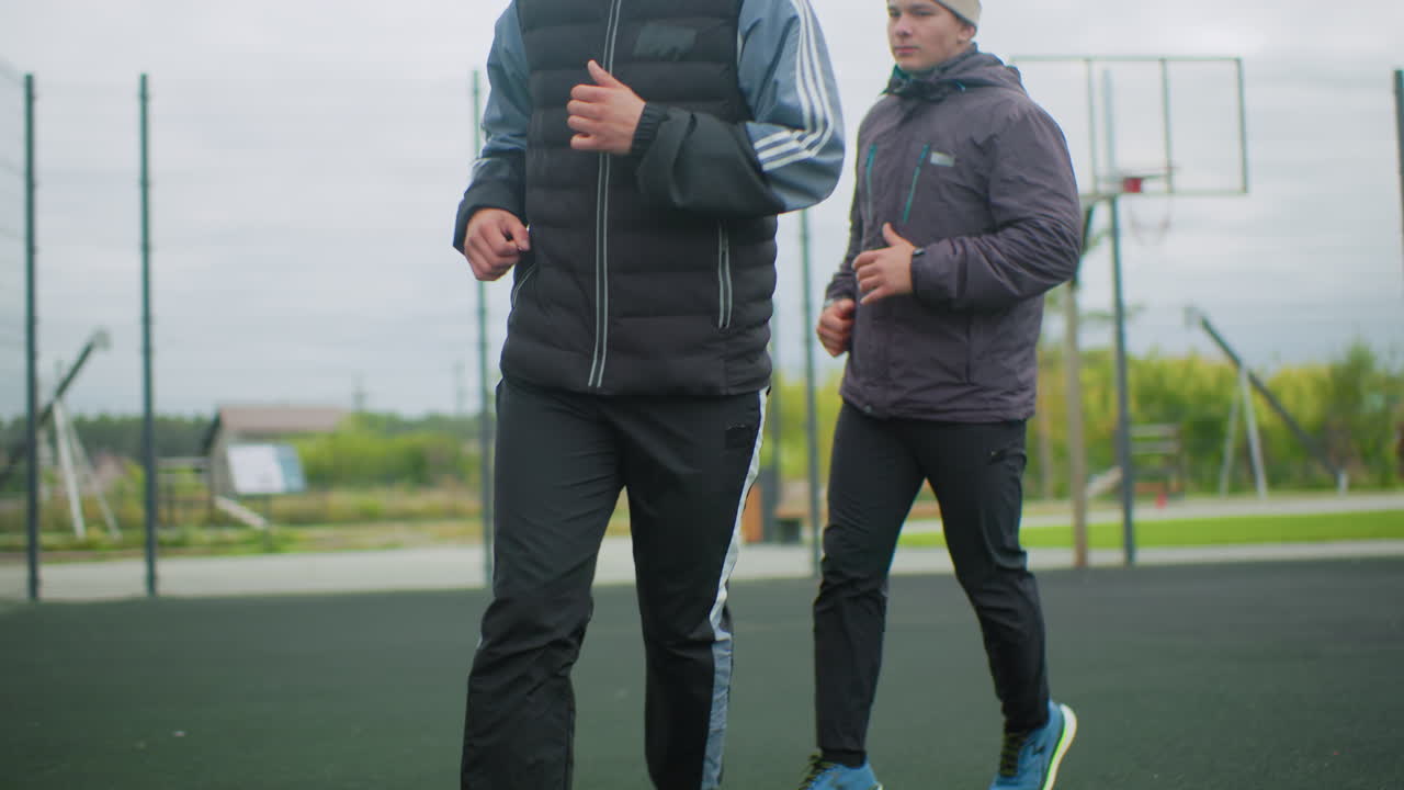 Due uomini bianchi che fanno jogging su un campo, indossando tute e scarpe da ginnastica, un allenatore che segue un giovane corridore durante una sessione mattutina nuvolosa, primi piani di passi su una corsia di asfalto con segnaletica gialla, pallacanestro