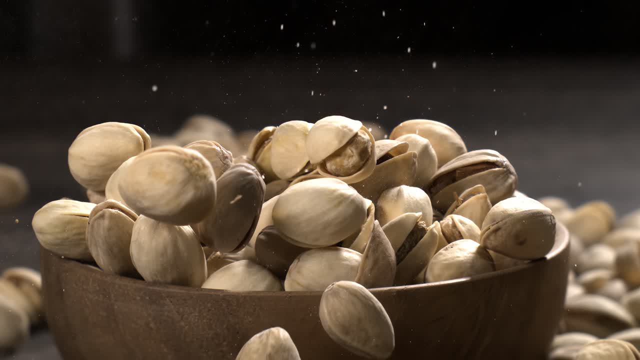 Ciotola di legno piena di pistacchi su tavolo di legno scuro, gustosa frutta secca salata sparsa intorno, concetto di snack salutare e cibo naturale. Scatto con telecamera super slow motion a 1600 fps