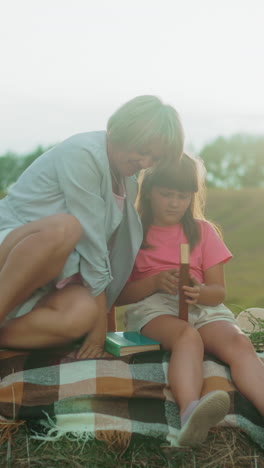 madre sentada con su hija pequeña en una pala de heno, compartiendo un momento juntos mientras la niña sostiene un libro de portada marrón, rodeada por un campo tranquilo