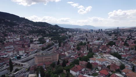 Aéreo:-Paisaje-Urbano-De-Sarajevo-Con-Río,-Edificios-Históricos-Y-Colinas