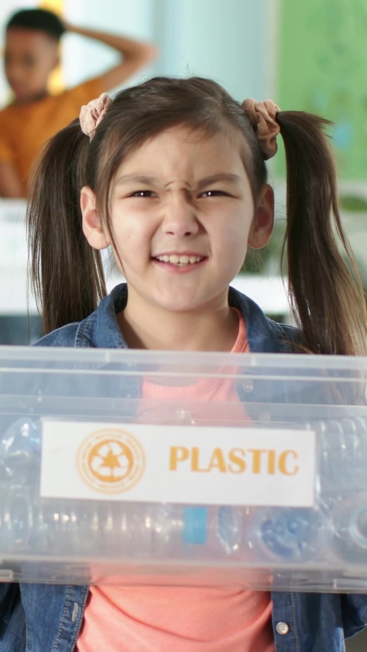 Video verticale: La telecamera si avvia, ragazza in denim che tiene un contenitore di PLASTICA con bottiglie in classe per il riciclo