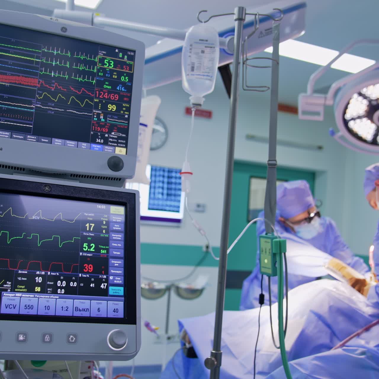 Monitore für Beatmungsgeräte und Tropfenzähler im Operationssaal. Zwei männliche Ärzte arbeiten an dem Operationstisch im Hintergrund.