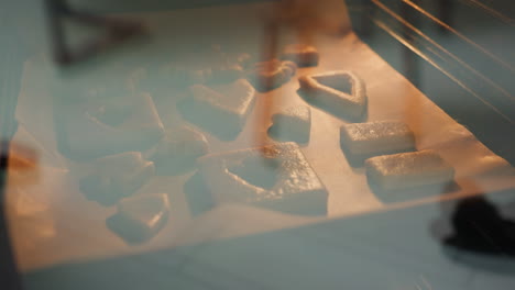 galletas doradas en diferentes formas horneando dentro de un horno visto a través del vidrio del horno, la cálida luz dorada del horno ilumina las galletas mientras hornean en papel de pergamino