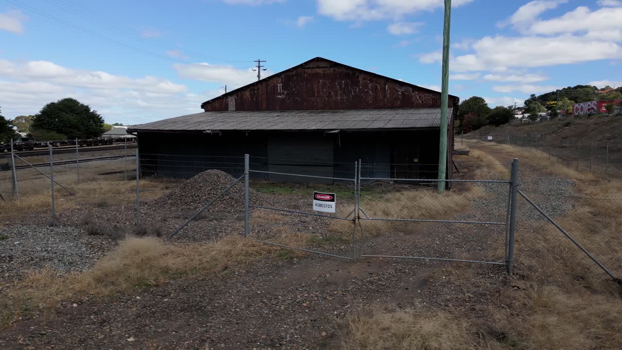An abandoned industrial shed with a corrugated roof and rusted exterior stands fenced off beside a railway line. A "Danger Asbestos" warning sign is displayed on the locked gate