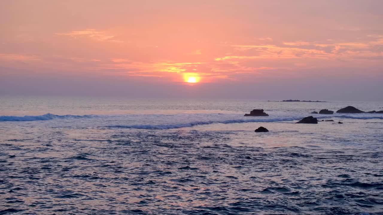 Overlooking rolling ocean waves during sunset with hazy pink orange sky in southern Sri Lanka
