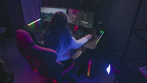 aerial view of woman with long hair wearing headphones focused on playing intense computer game using dual monitors and microphone in dark room lit with purple ambient lights and professional gear