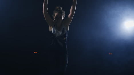 a young female gymnast runs up looking at the camera and performs a forward flip and screw in the air in slow motion in the gym on the carpet in the smoke. the steadicam in motion
