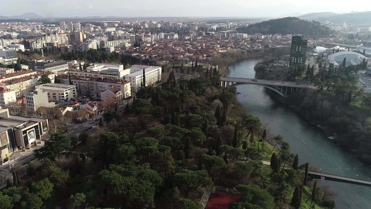 Ripresa aerea della città di Podgorica verso i pini. Giornata soleggiata e luminosa con un cielo blu.