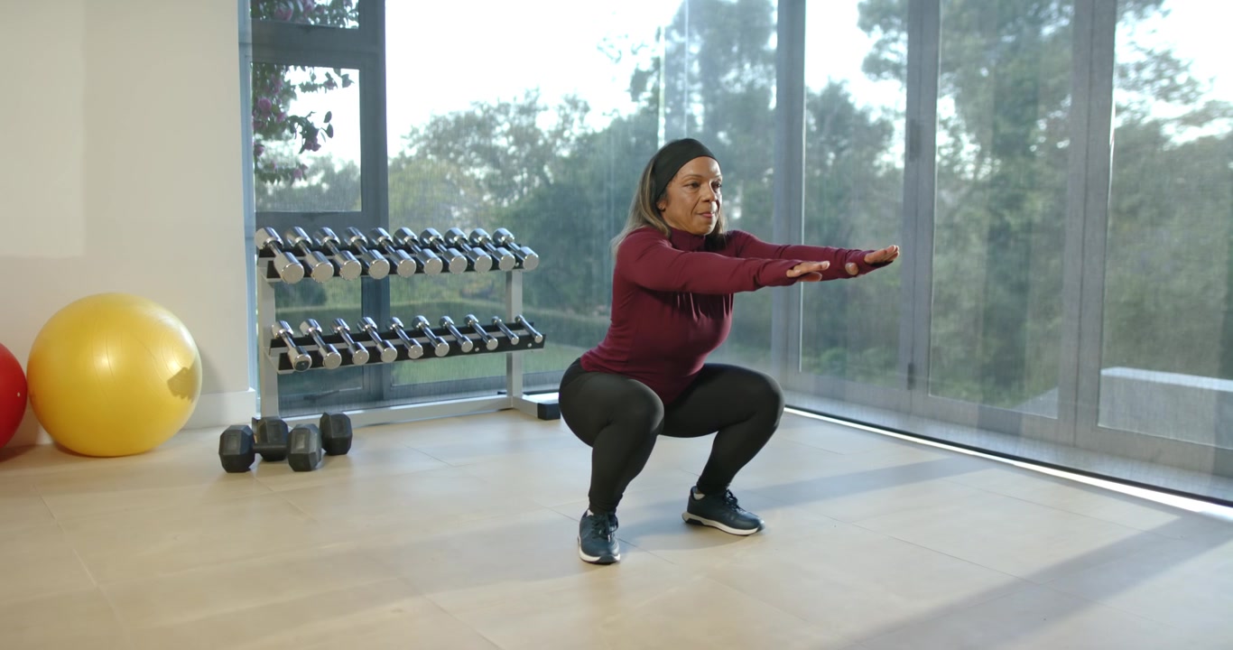 Donna afroamericana matura che fa squat spingendo indietro i fianchi, facendo allenamento a casa vicino a una rastrelliera per manubri.