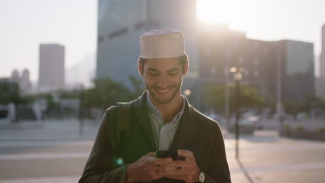 close up portrait of attractive young muslim man texting browsing using smartphone mobile messaging app on urban city street at sunset