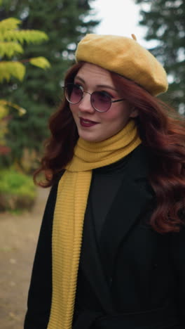 artista con el cabello rojo largo ondulado, con un abrigo negro y boina amarilla, ajusta su cabello mientras camina a través de un parque exuberante, rodeado de colorido follaje de otoño