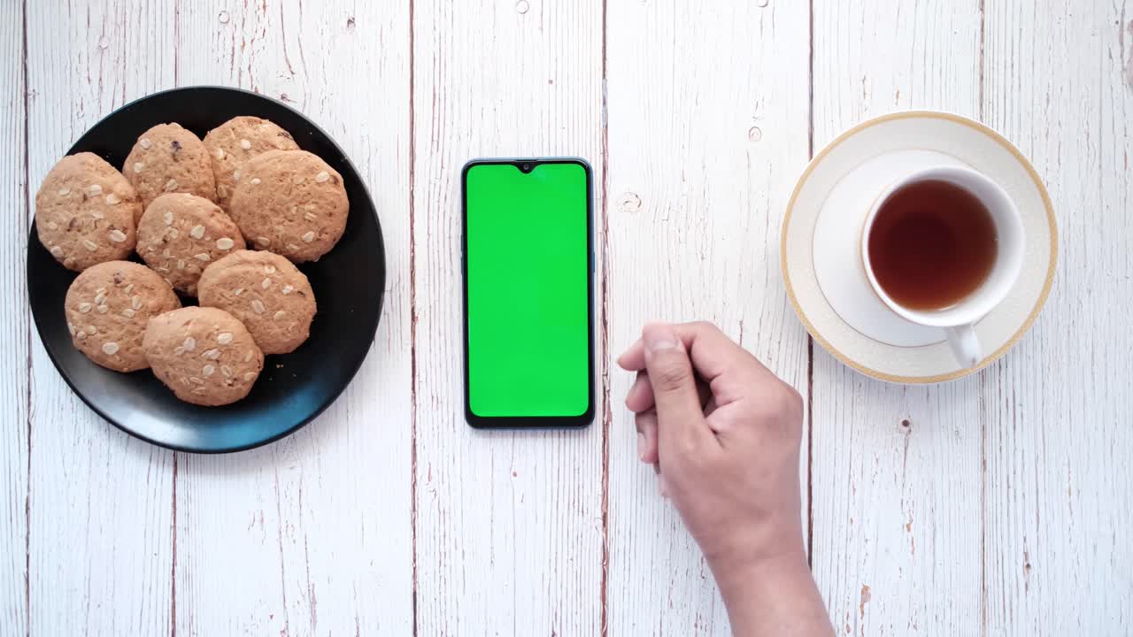 teléfono inteligente con pantalla verde, galletas y té en una mesa de madera