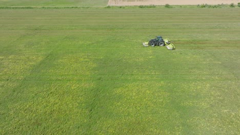 Aerial-establishing-view-of-a-tractor-mowing-a-fresh-green-grass-field,-a-farmer-in-a-modern-tractor-preparing-food-for-farm-animals,-sunny-summer-day,-wide-drone-dolly-shot-moving-left