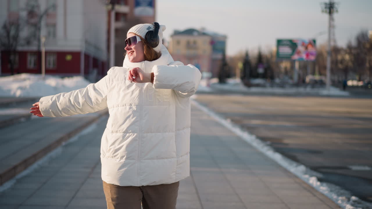 Young woman gliding along city sidewalk with arms raised, moving to rhythm in winter attire, wearing headset and sunglasses, joyful expression while crossing urban street with snow piles