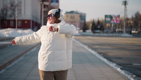 young woman gliding along city sidewalk with arms raised, moving to rhythm in winter attire, wearing headset and sunglasses, joyful expression while crossing urban street with snow piles