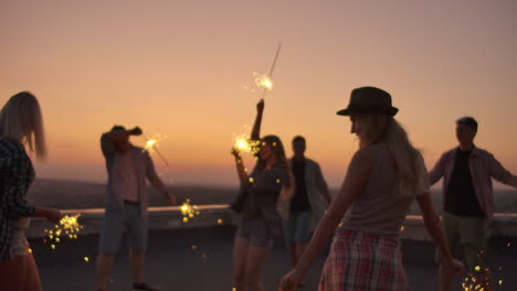 The-company-of-young-people-moves-their-hands-and-body-on-the-roof-with-big-bengal-light-in-summer-evening.-They-celebrate-the-birth-of-a-friend.