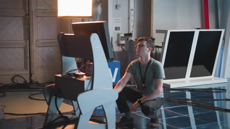 man crouching on floor in studio, adjusting monitors and equipment, preparing for video production or broadcast, illuminated by overhead lights with cables and equipment visible in background