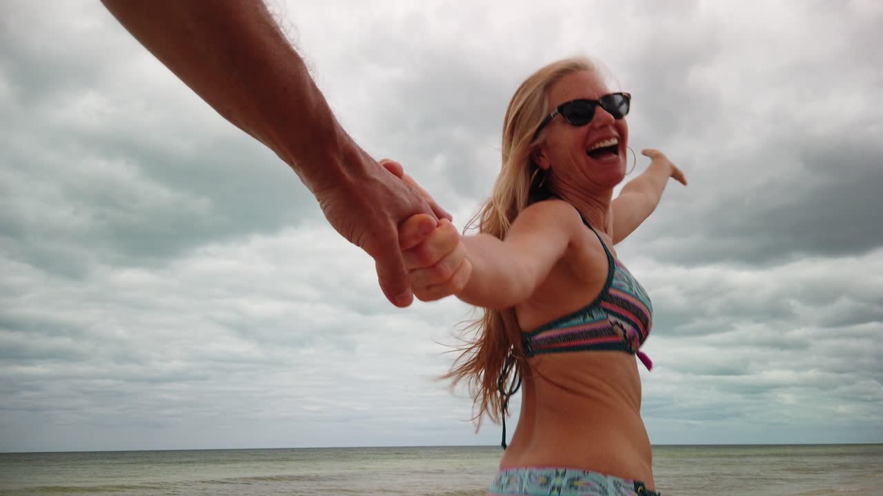 Looking back a mature woman run bikini and sunglasses running in slow motion in follow me pulling her husband on the beach